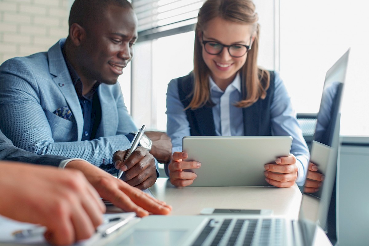 Executive woman in glasses and smiling young african american man in blazer reviewing a document and working on a laptop