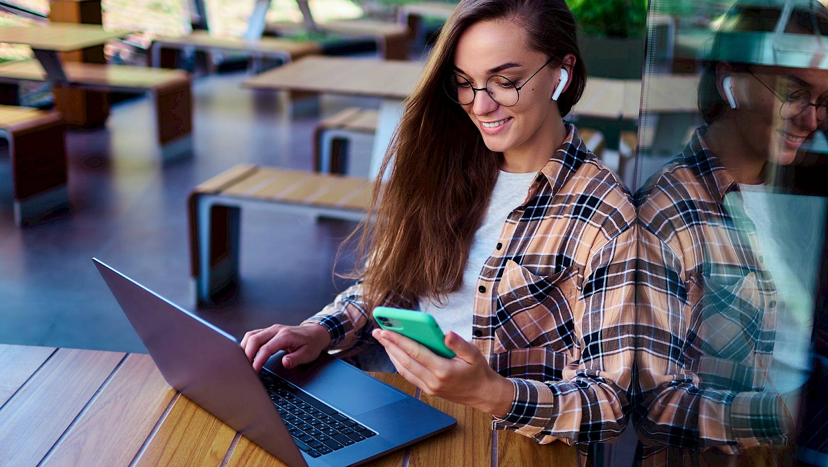 Young female smiling wearing ear pods holding a smartphone and using a laptop