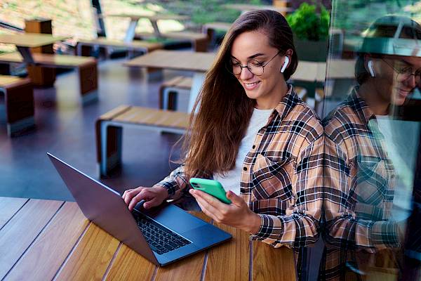 Young female smiling wearing ear pods holding a smartphone and using a laptop